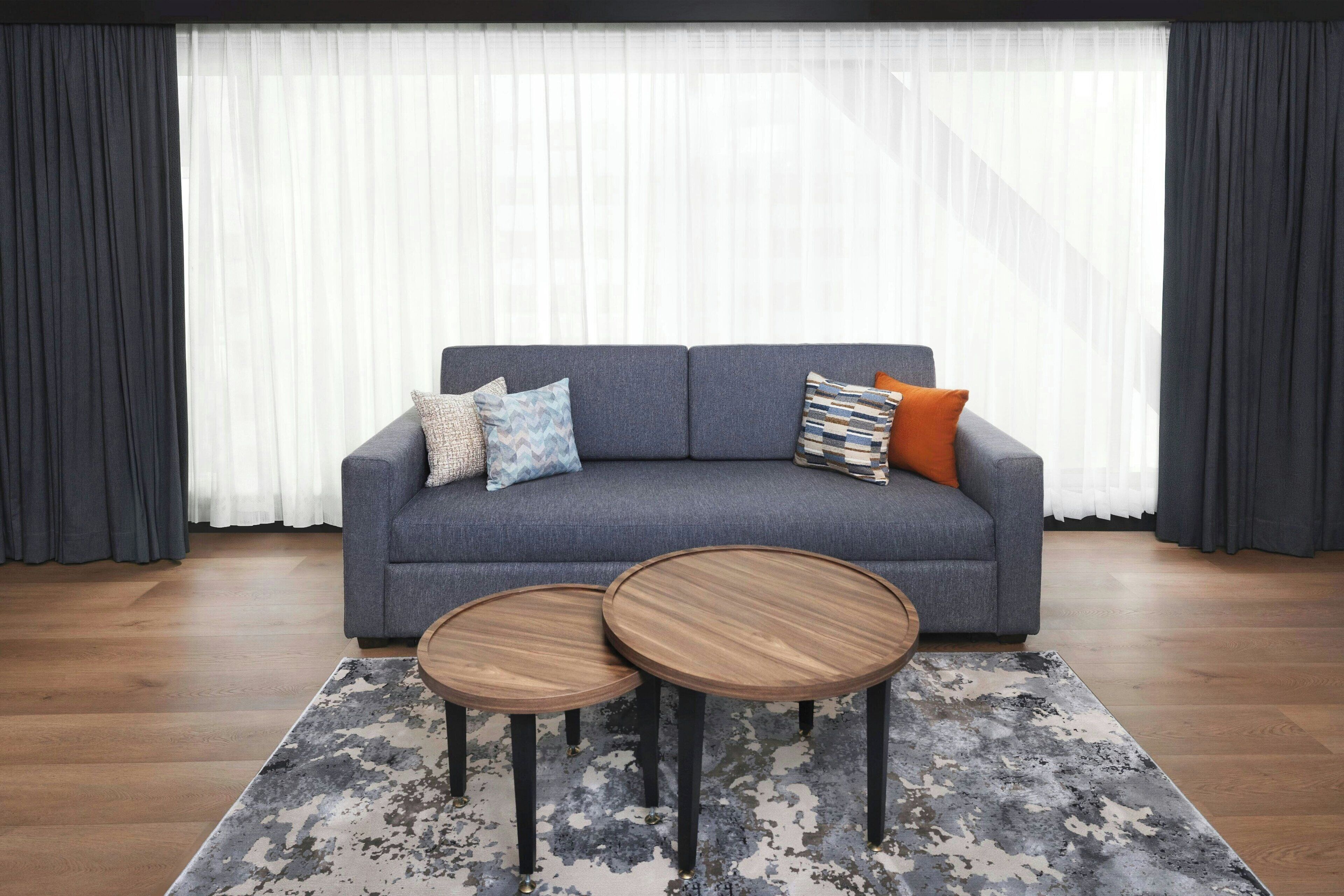 A grey sofa with patterned cushions, two round wooden side tables, and a grey and white abstract rug.