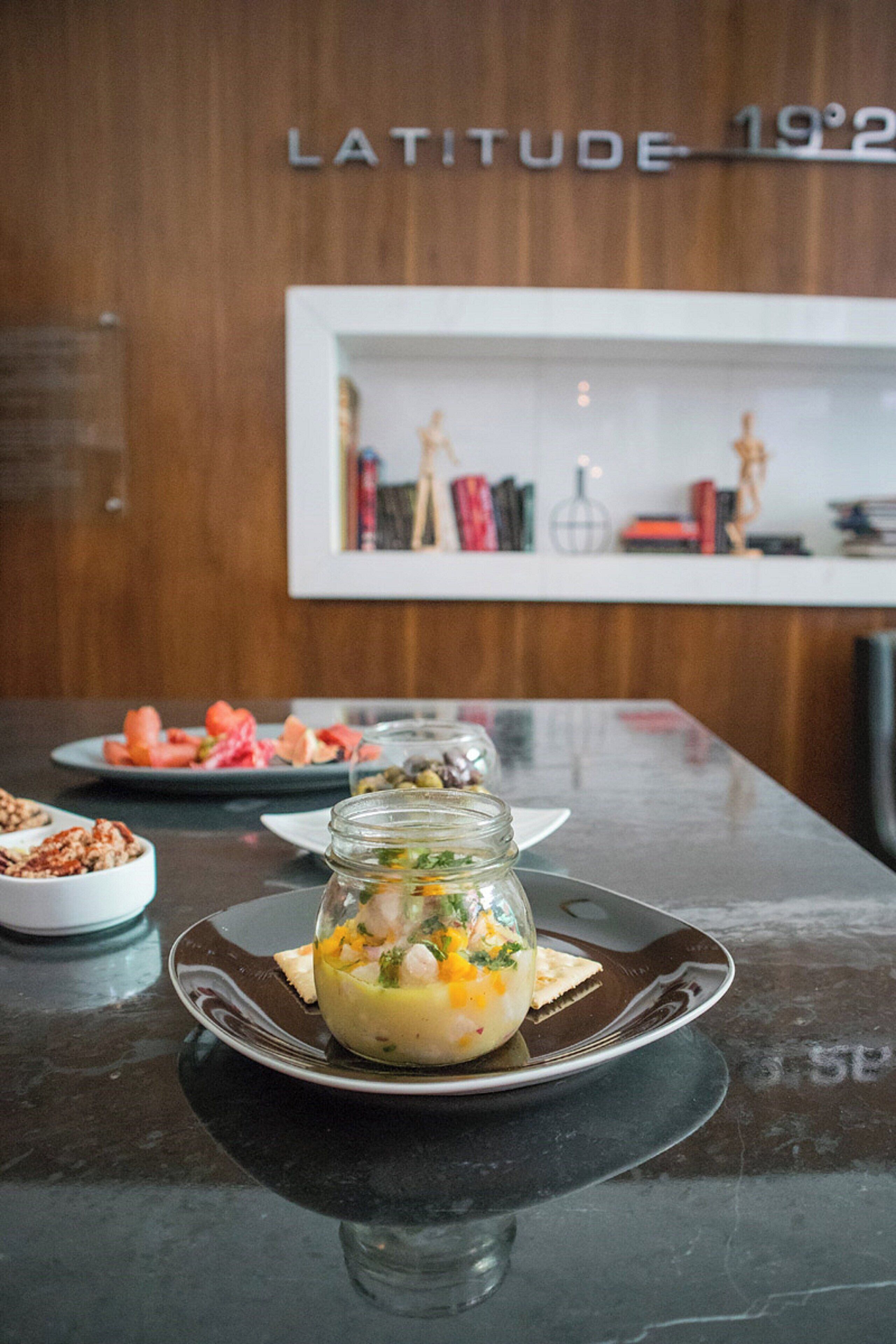 Une table à manger avec une assiette de nourriture, un bocal de salade et un dessert. Au fond, on aperçoit une étagère remplie de livres et une pancarte indiquant « LATITUDE 19°2 ».