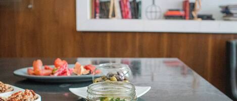 Une table à manger avec une assiette de nourriture, un bocal de salade et un dessert. Au fond, on aperçoit une étagère remplie de livres et une pancarte indiquant « LATITUDE 19°2 ».