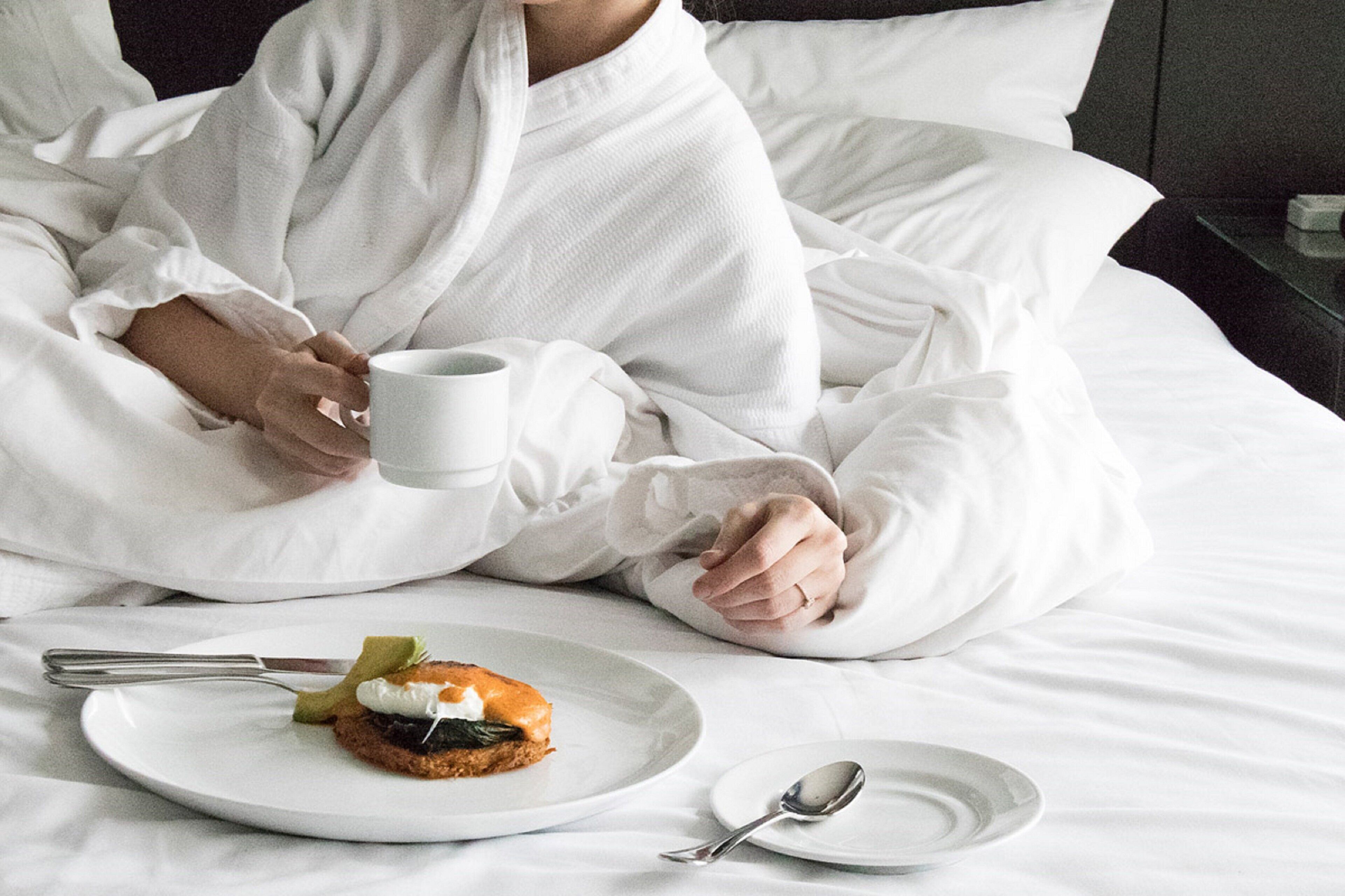 A person in a white robe sitting on a bed with a plate of food and a mug.
