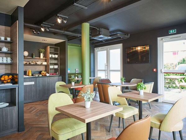 A modern cafe interior with wooden tables and chairs, a bar area with shelves, and a large window providing natural light. - Kyriad Rouen Sud - Val De Reuil (Val-de-Reuil)