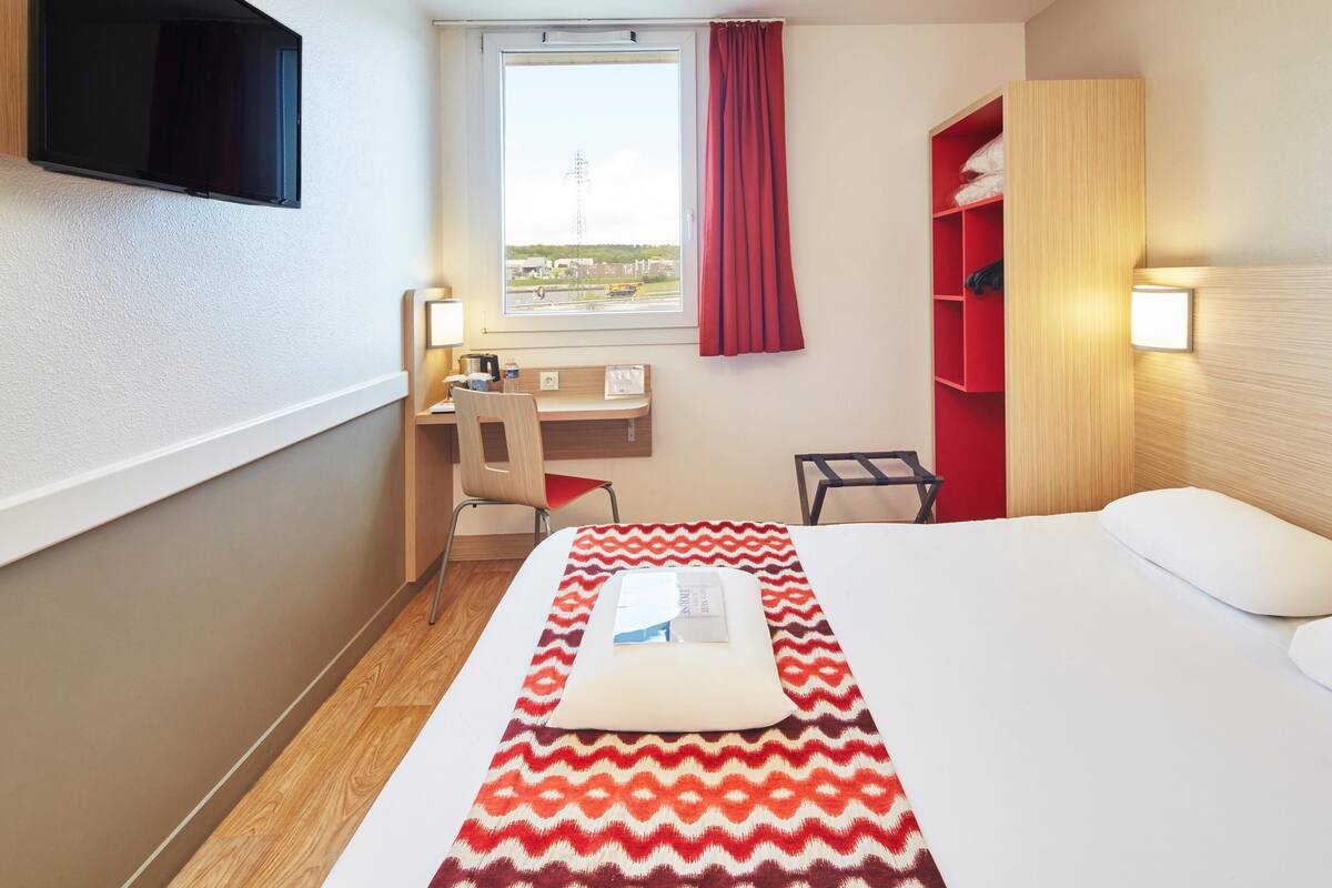 A hotel room with a bed, a desk with a chair, a TV mounted on the wall, a window with a curtain, and a shelf with towels.