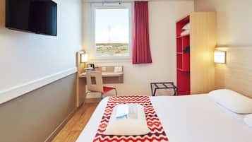 A hotel room with a bed, a desk with a chair, a TV mounted on the wall, a window with a curtain, and a shelf with towels.