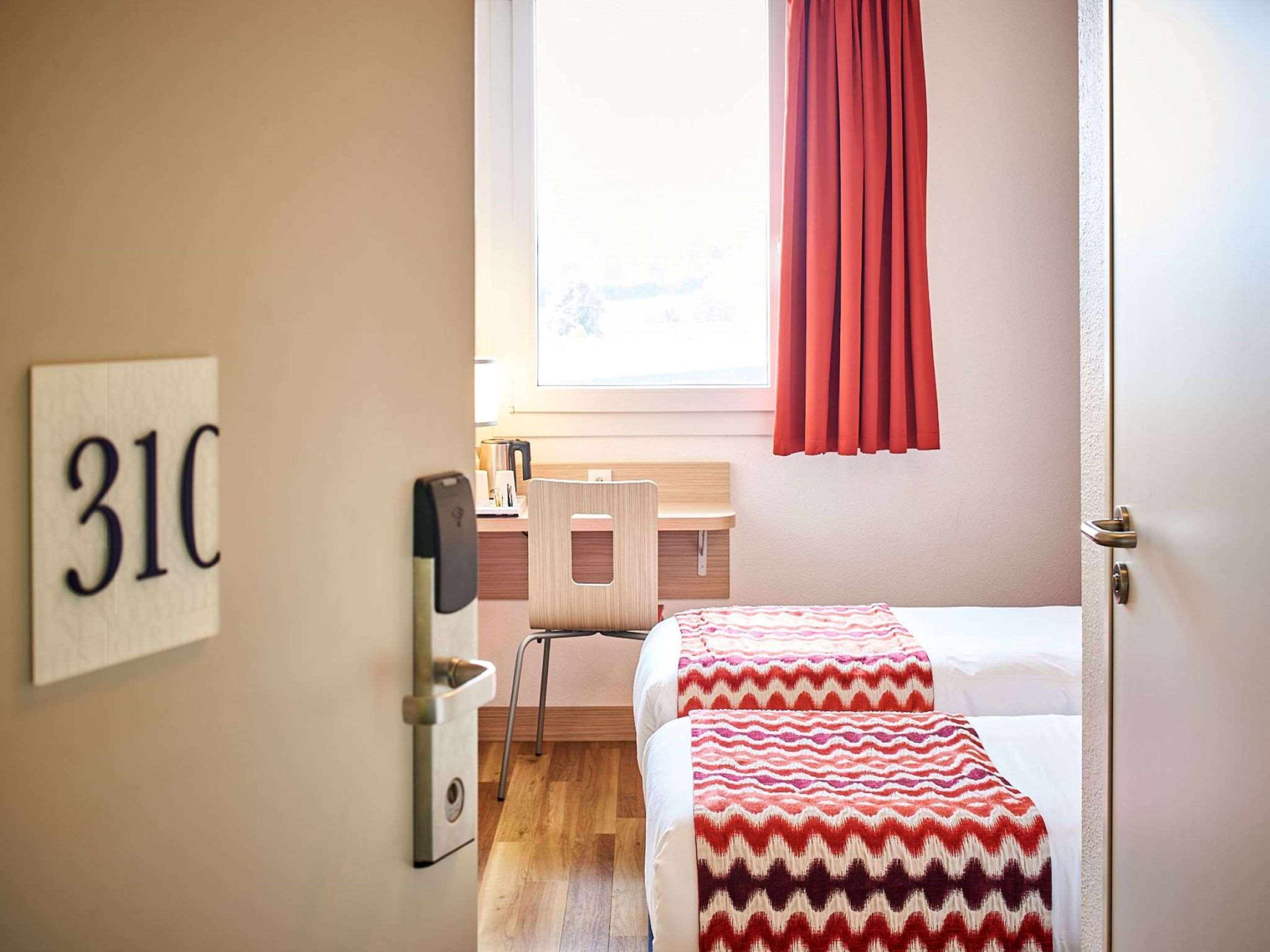 A hotel room with two beds, a desk, a chair, a window with red curtains, and a wall-mounted number sign.