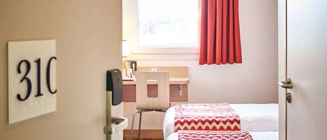A hotel room with two beds, a desk, a chair, a window with red curtains, and a wall-mounted number sign.