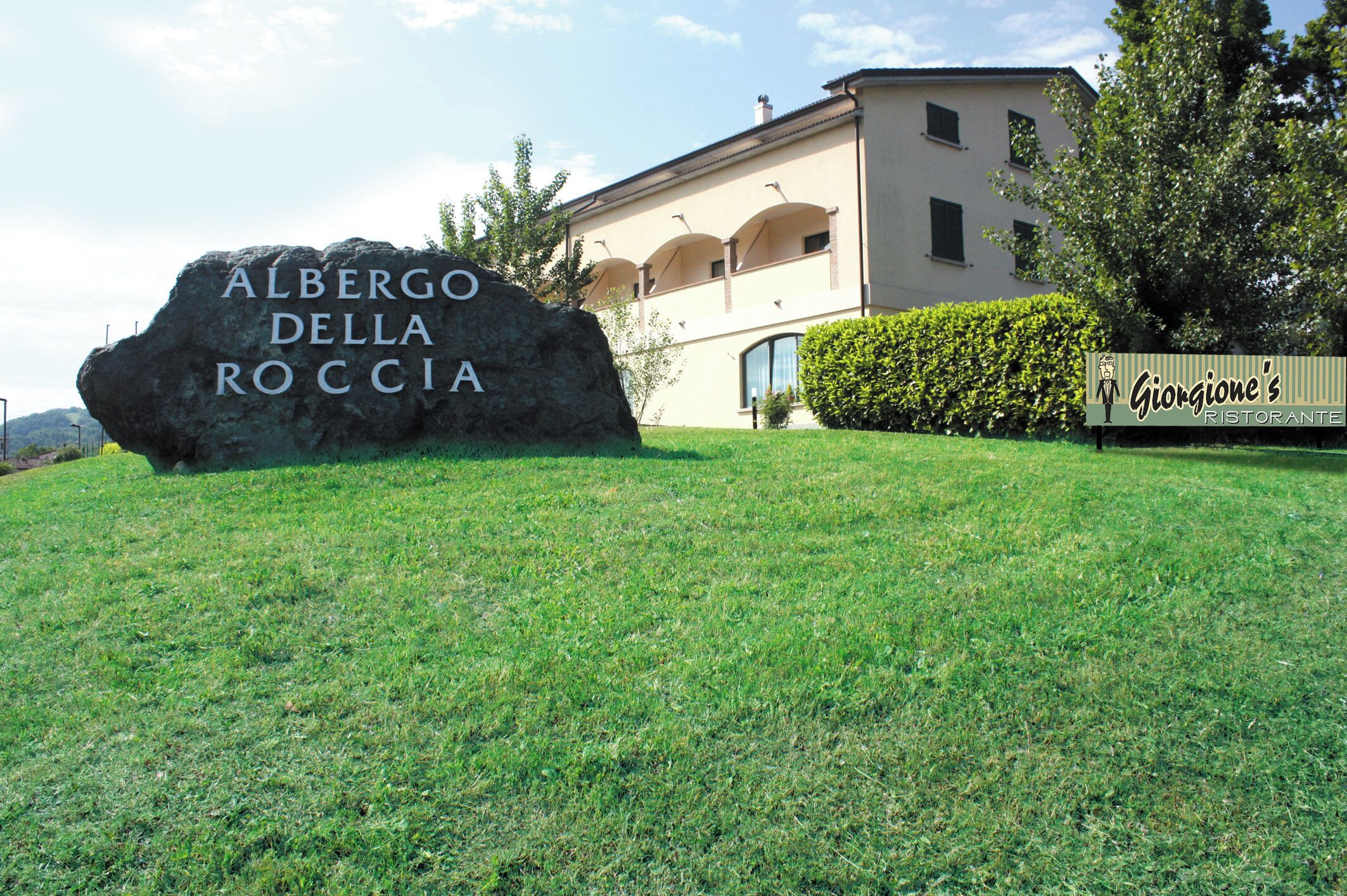 Un grand rocher sur lequel est gravé le nom « Albergo della Roccia », un bâtiment à la façade beige et aux fenêtres en arc de cercle, et une enseigne indiquant « Giorgione's Ristorante » à l’avant.