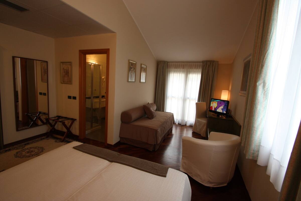 Quarto de hotel com uma cama grande, um espaço de estar com uma poltrona, televisão e escrivaninha.