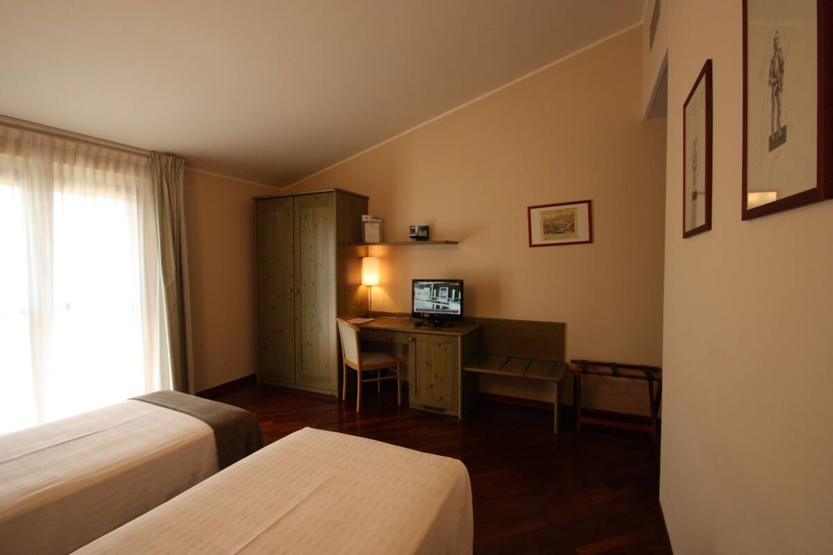 Quarto de hotel com duas camas, uma escrivaninha com computador, uma cadeira, um guarda-roupa e uma pequena prateleira com um quadro.