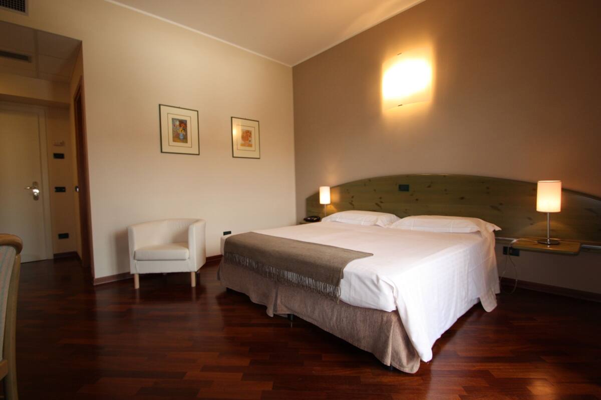 Quarto de hotel com uma cama grande, uma cadeira e duas fotos emolduradas na parede.