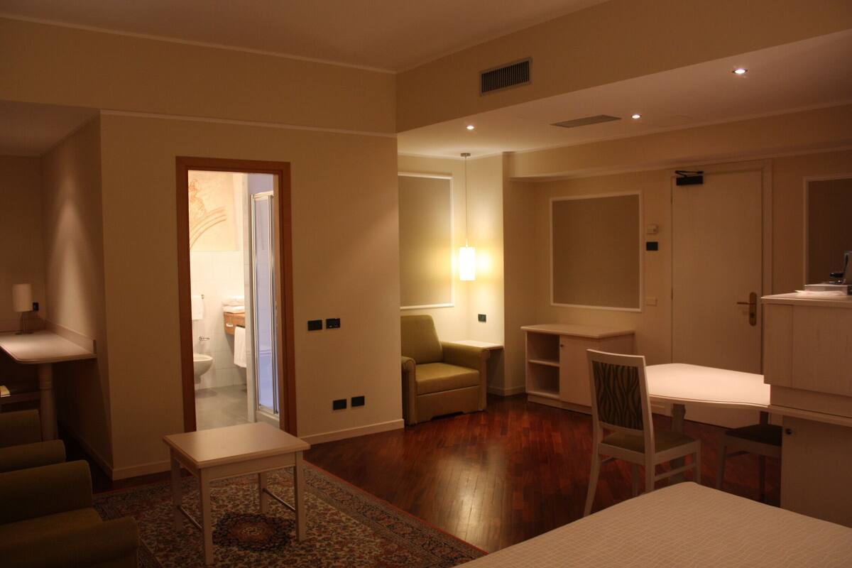 Quarto de hotel com cama, escrivaninha, cadeira, mesinha, sofá e banheiro visível através de uma porta aberta.