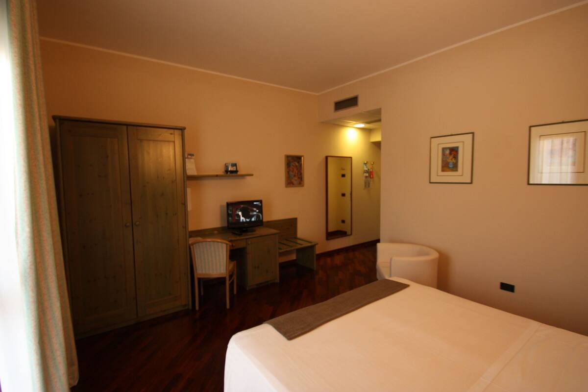Quarto de hotel com cama, escrivaninha, televisão, cadeira, armário e banheiro.