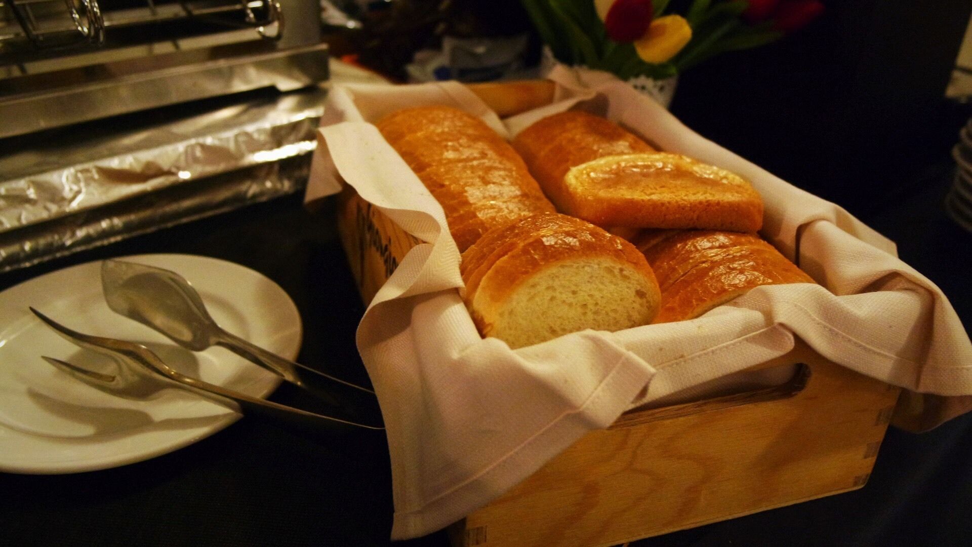 Un cesto di legno con diversi filoni di pane, un piatto con un cucchiaio e un vaso con fiori.