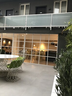 Uma área de refeições ao ar livre com mesa e cadeiras brancas, guarda-corpo de vidro e vista para o espaço interior.