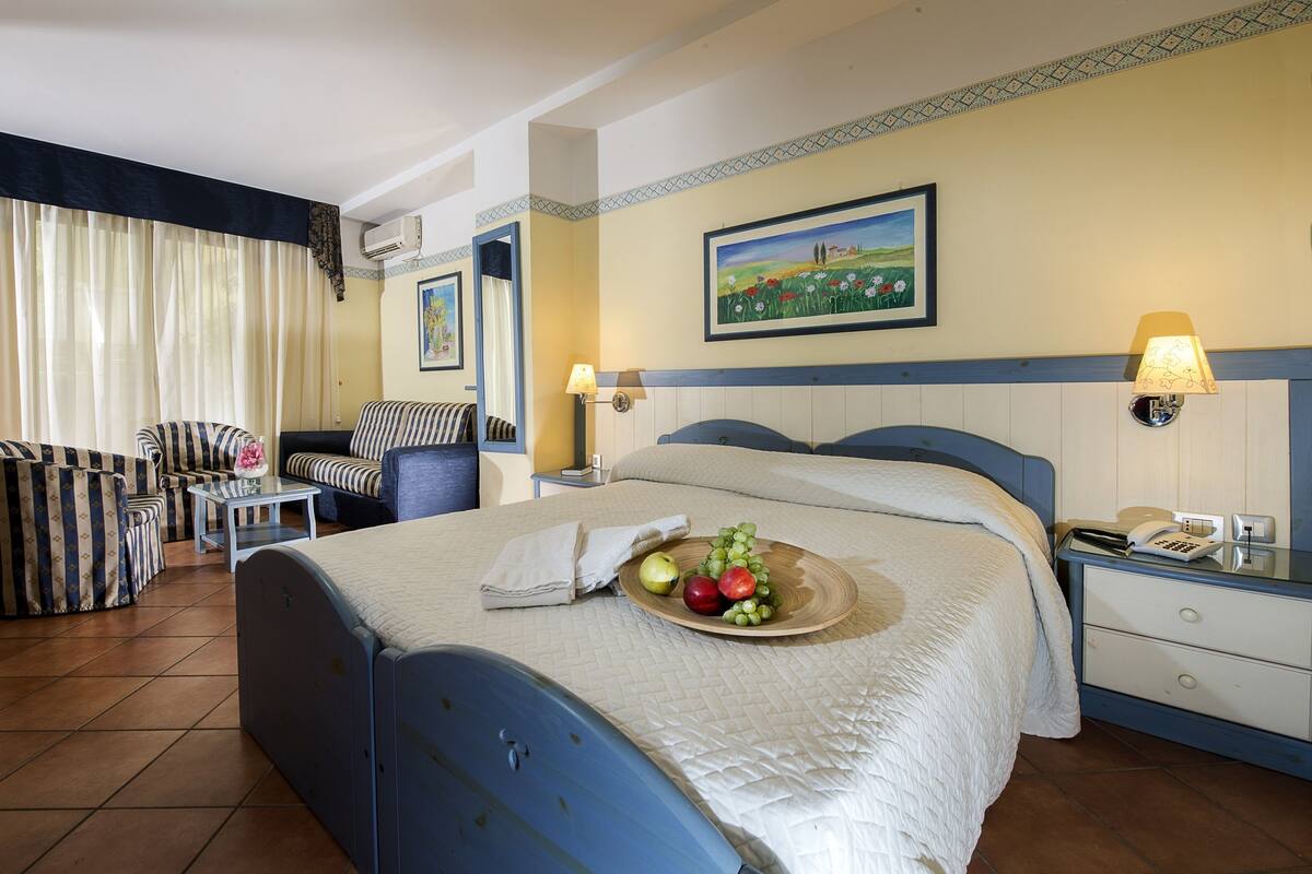 A hotel room with a bed, a chair, a small table, a lamp, and a painting on the wall.