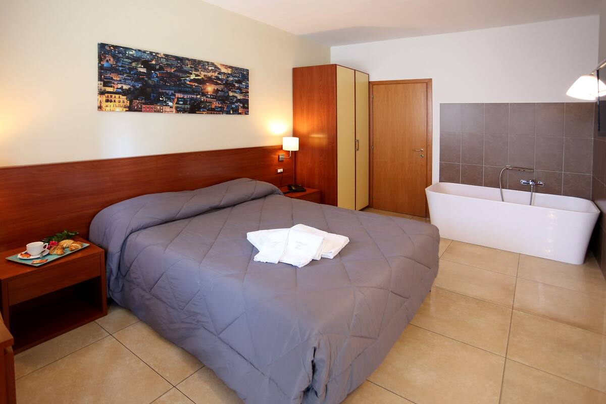 A hotel room with a large bed, a wooden headboard, a nightstand with a lamp, a painting on the wall, a wardrobe, and a bathtub.
