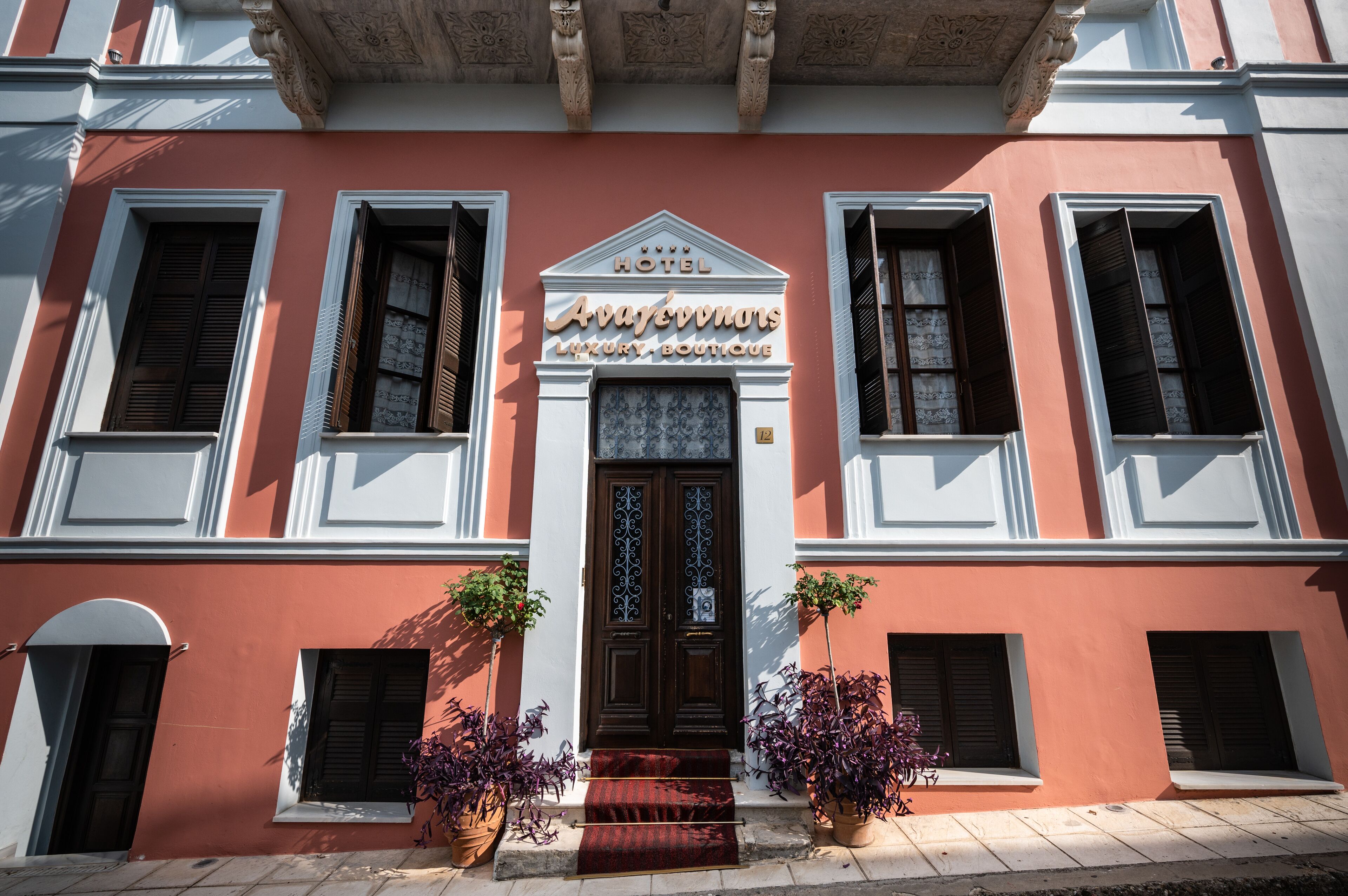 A hotel facade with a central entrance, flanked by windows with shutters, and potted plants at the entrance.