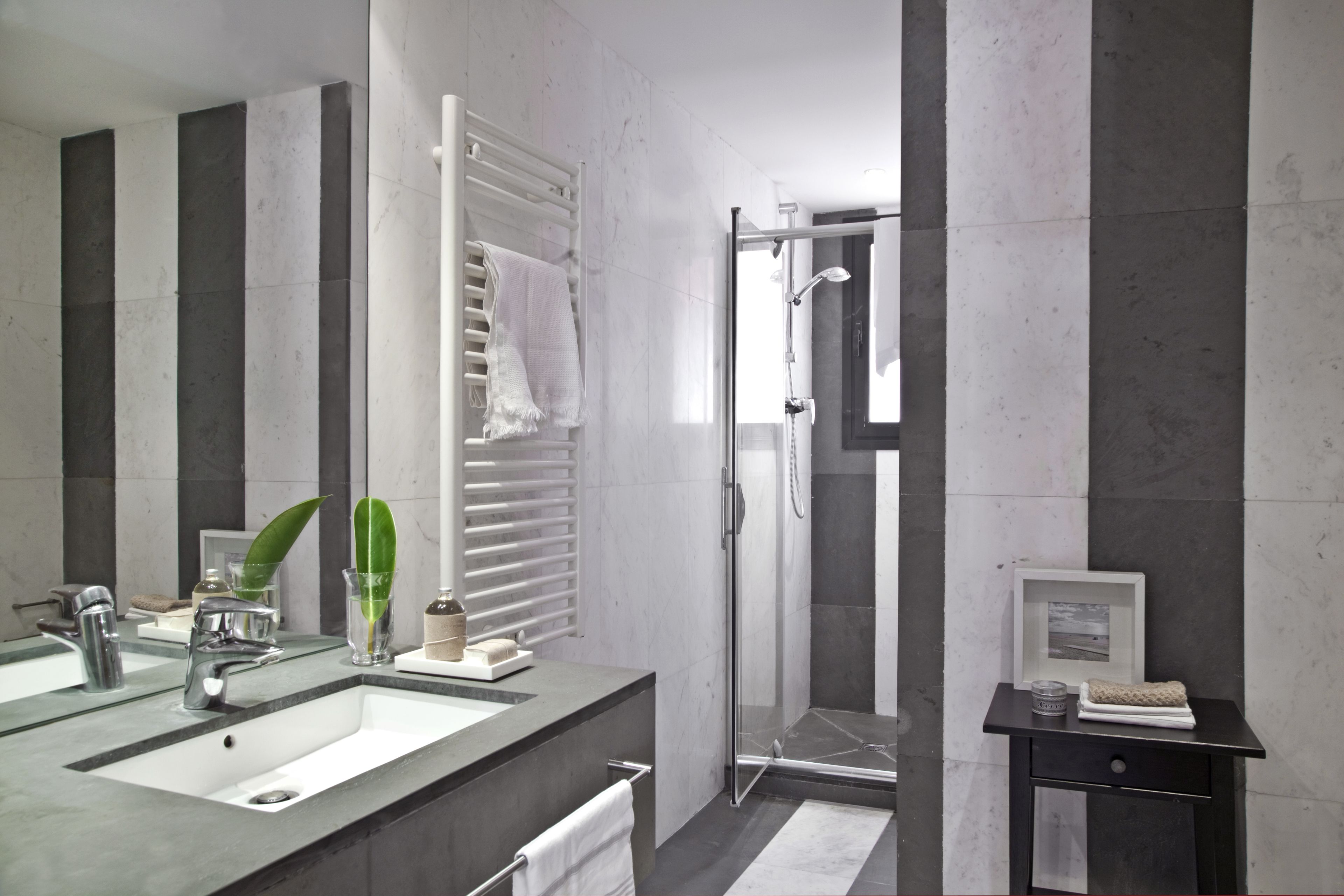 Een moderne badkamer met een grote spiegel, twee wasbakken, een douche en een klein nachtkastje met een afbeelding in een lijst.