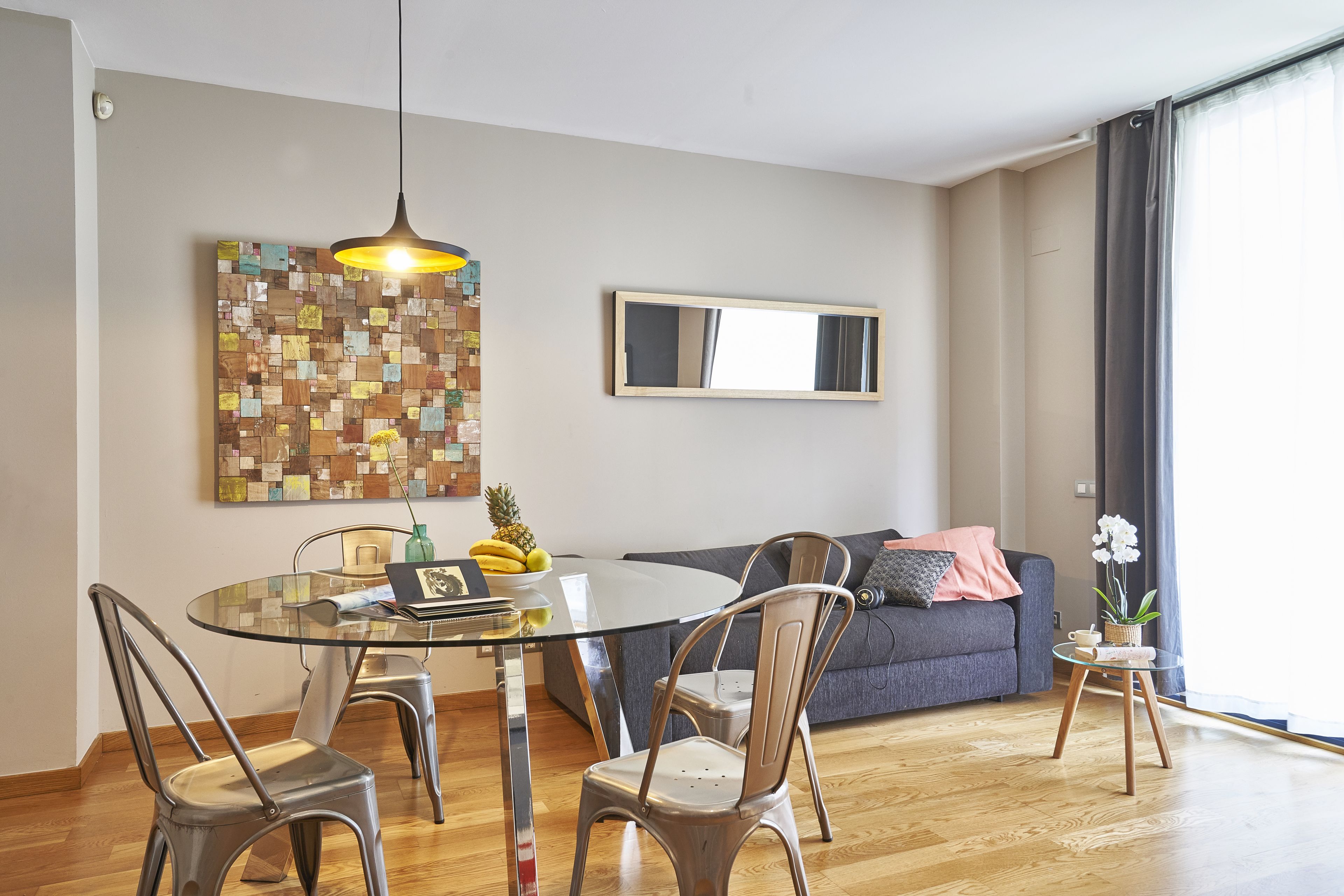Una sala moderna con una mesa de comedor de vidrio, sillas de metal, un sofá gris, una pequeña mesita de noche con una planta y una llamativa obra de arte abstracto en la pared.