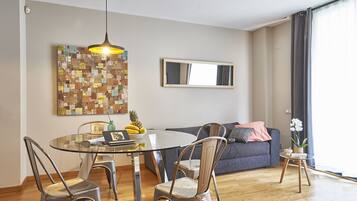 Una sala moderna con una mesa de comedor de vidrio, sillas de metal, un sofá gris, una pequeña mesita de noche con una planta y una llamativa obra de arte abstracto en la pared.