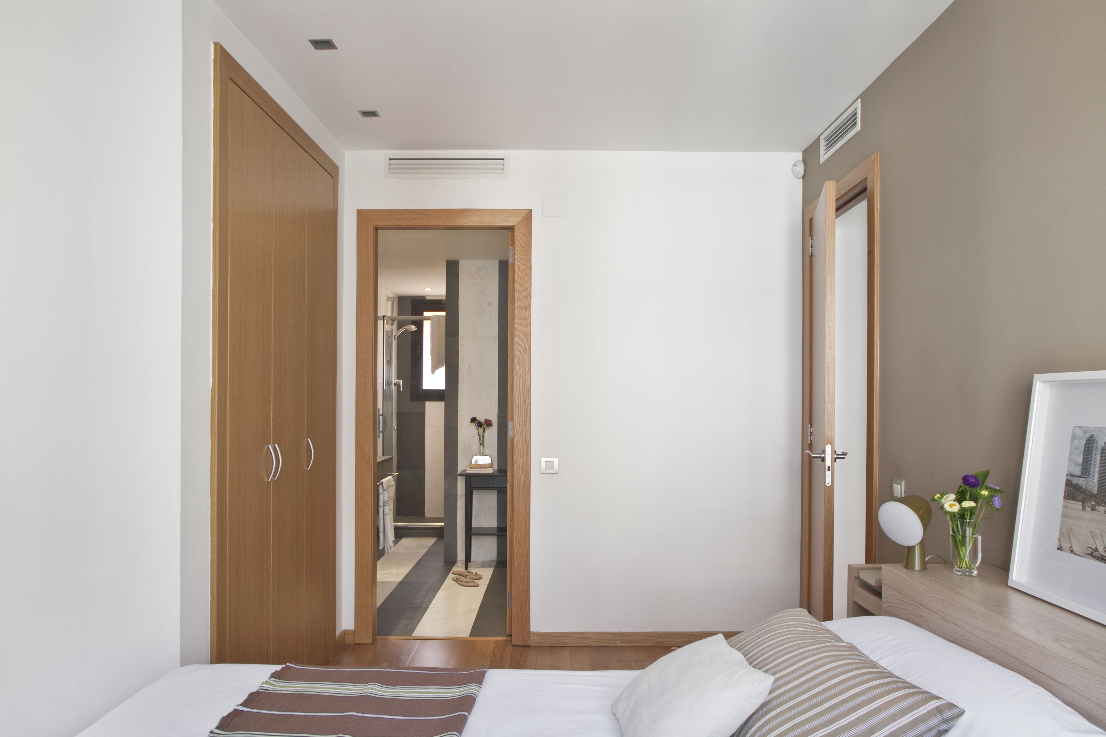 A bedroom with a bed, wooden wardrobe, a bedside table with a vase of flowers, and a framed picture on the wall.