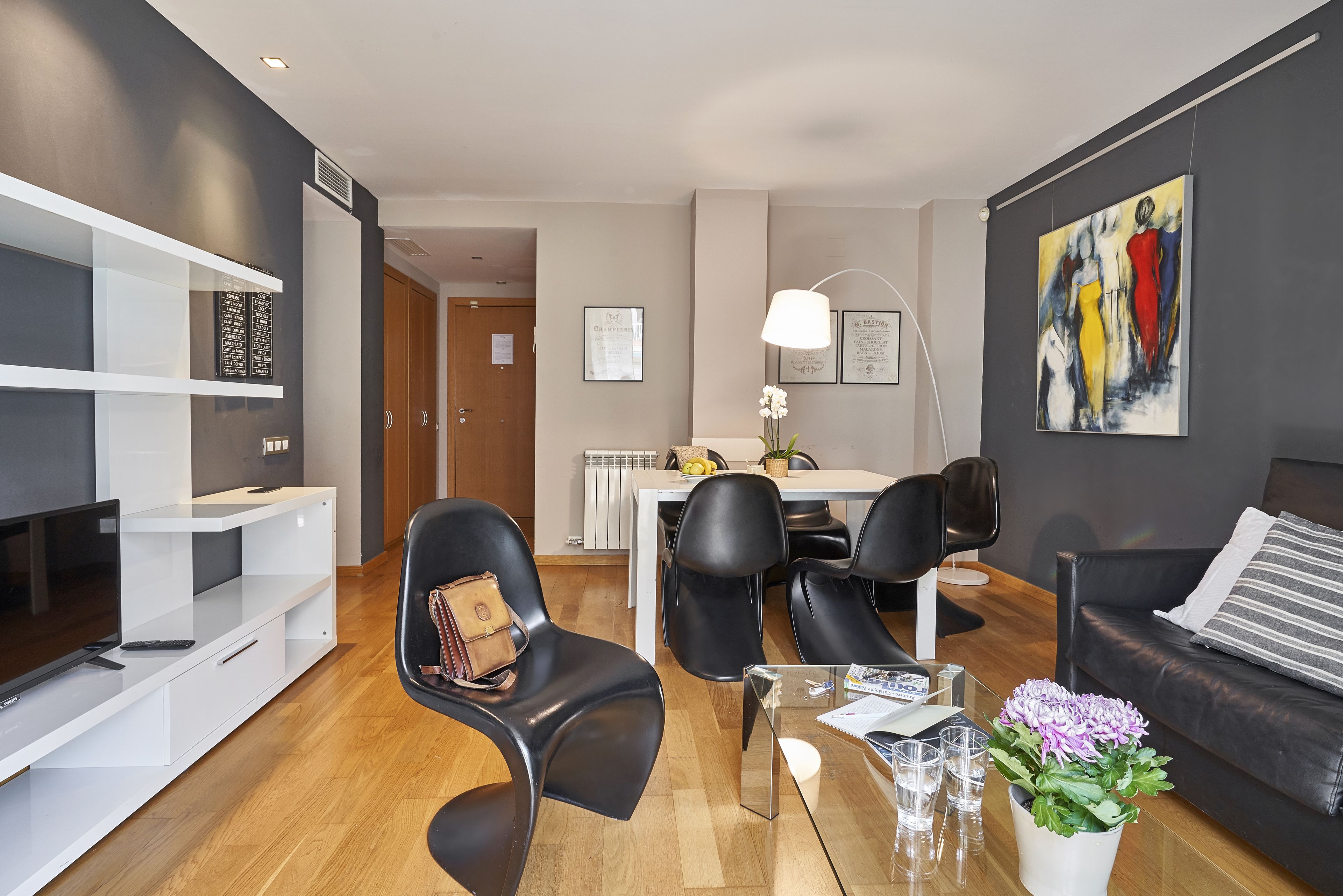 A modern living room with a flat-screen TV, black leather chairs, a glass coffee table, and a dining area with a white table and chairs.