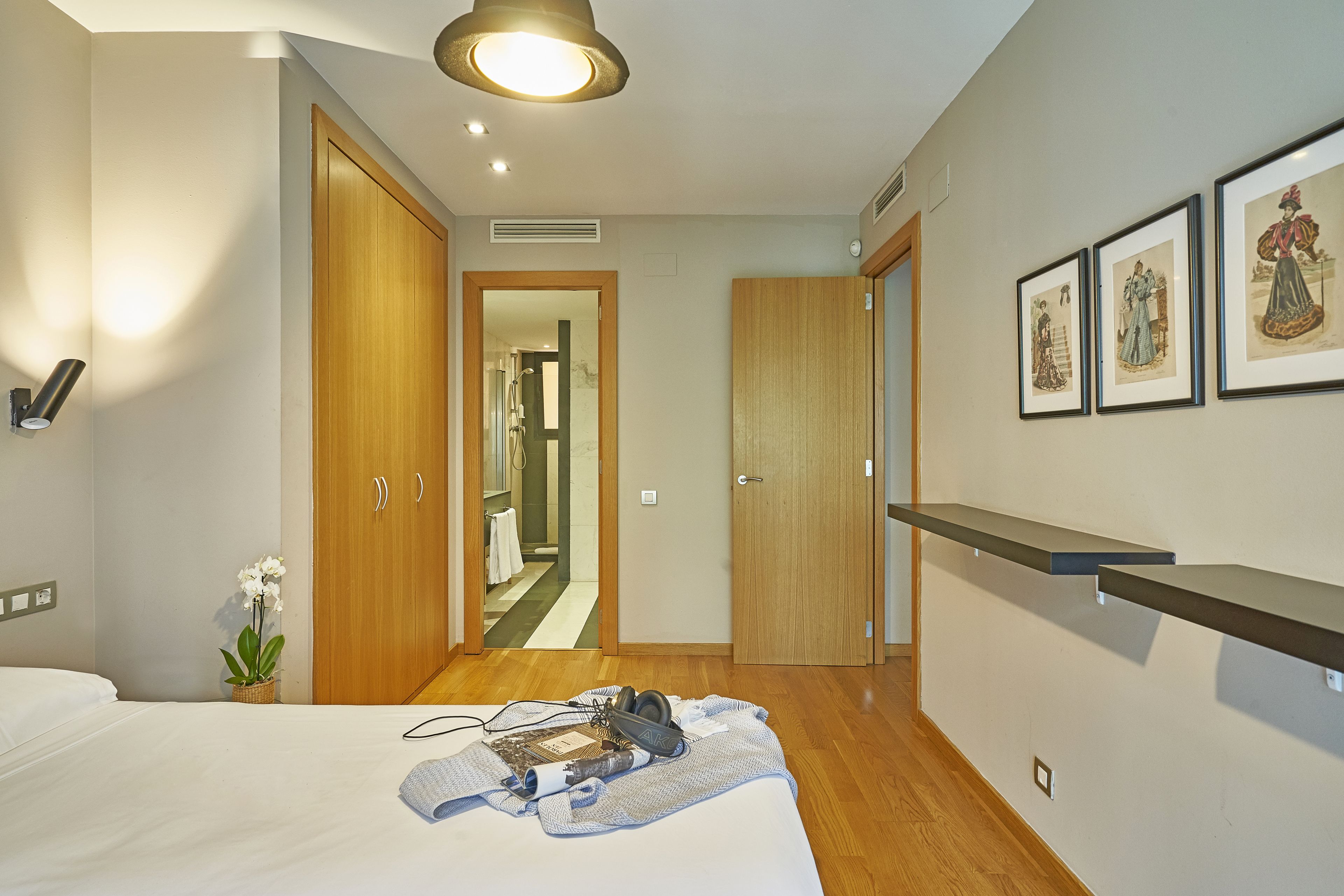 A hotel room with a bed, a bedside table with a lamp, a wall-mounted shelf, and framed artwork.
