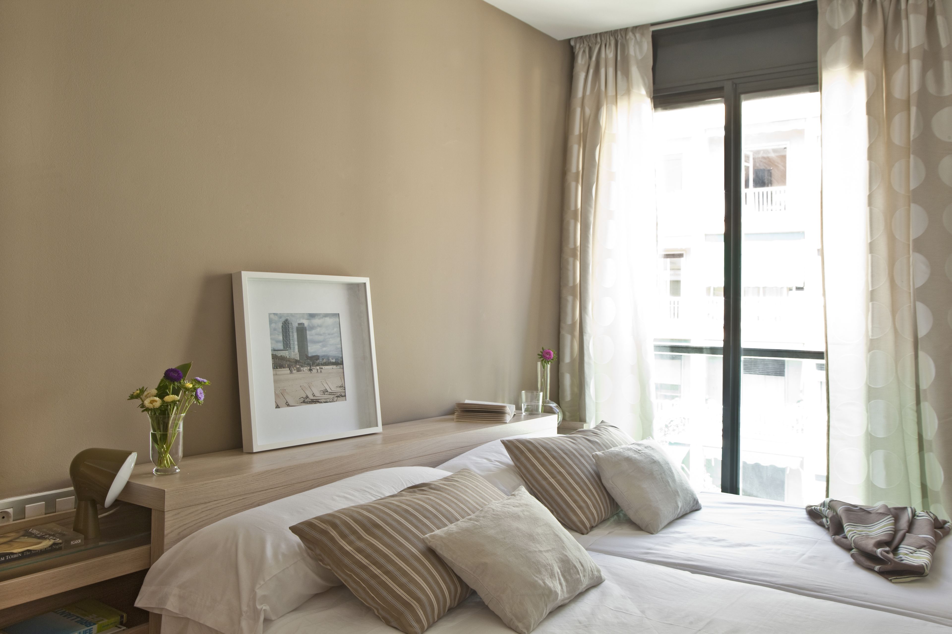 A hotel room with two beds, a desk with a lamp, a framed picture, a vase with flowers, and a window with curtains.