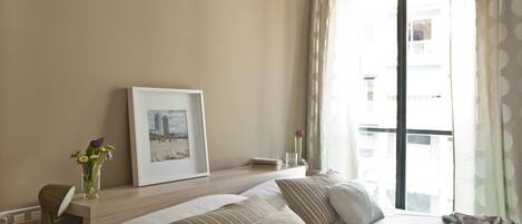 A hotel room with two beds, a desk with a lamp, a framed picture, a vase with flowers, and a window with curtains.