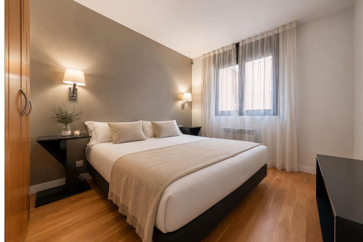 Een moderne slaapkamer met een groot bed, bedlampen, een nachtkastje, een raam met dunne gordijnen en een houten vloer.