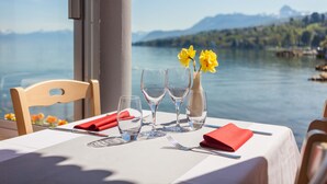 Mesa posta com taças de vinho, um vaso com flores amarelas e um guardanapo vermelho, com vista para um lago e montanhas ao fundo.