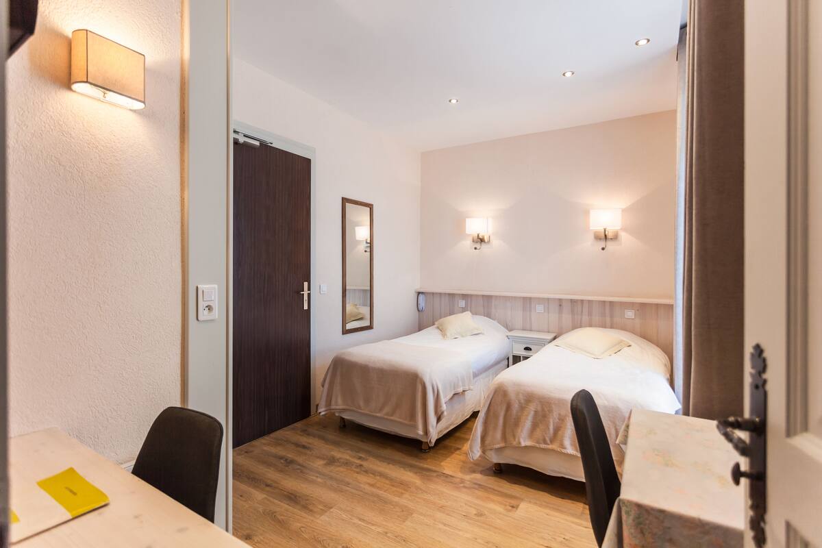 Habitación de hotel con dos camas, suelo de madera, un escritorio con silla e iluminación en la pared.
