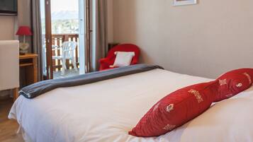 A hotel room with a bed, a red chair, a desk with a lamp, and a view of the outdoors.