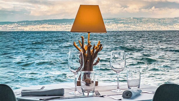 Mesa posta para refeição, com vista para o mar e paisagem urbana ao fundo.