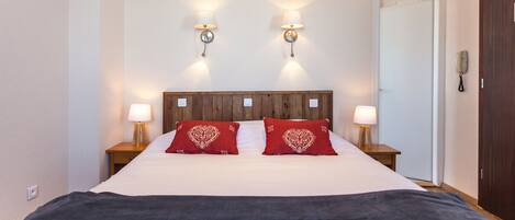 Habitación de hotel con cabecera de madera, dos lámparas de noche, una colcha blanca y dos almohadas rojas con estampado de corazones.