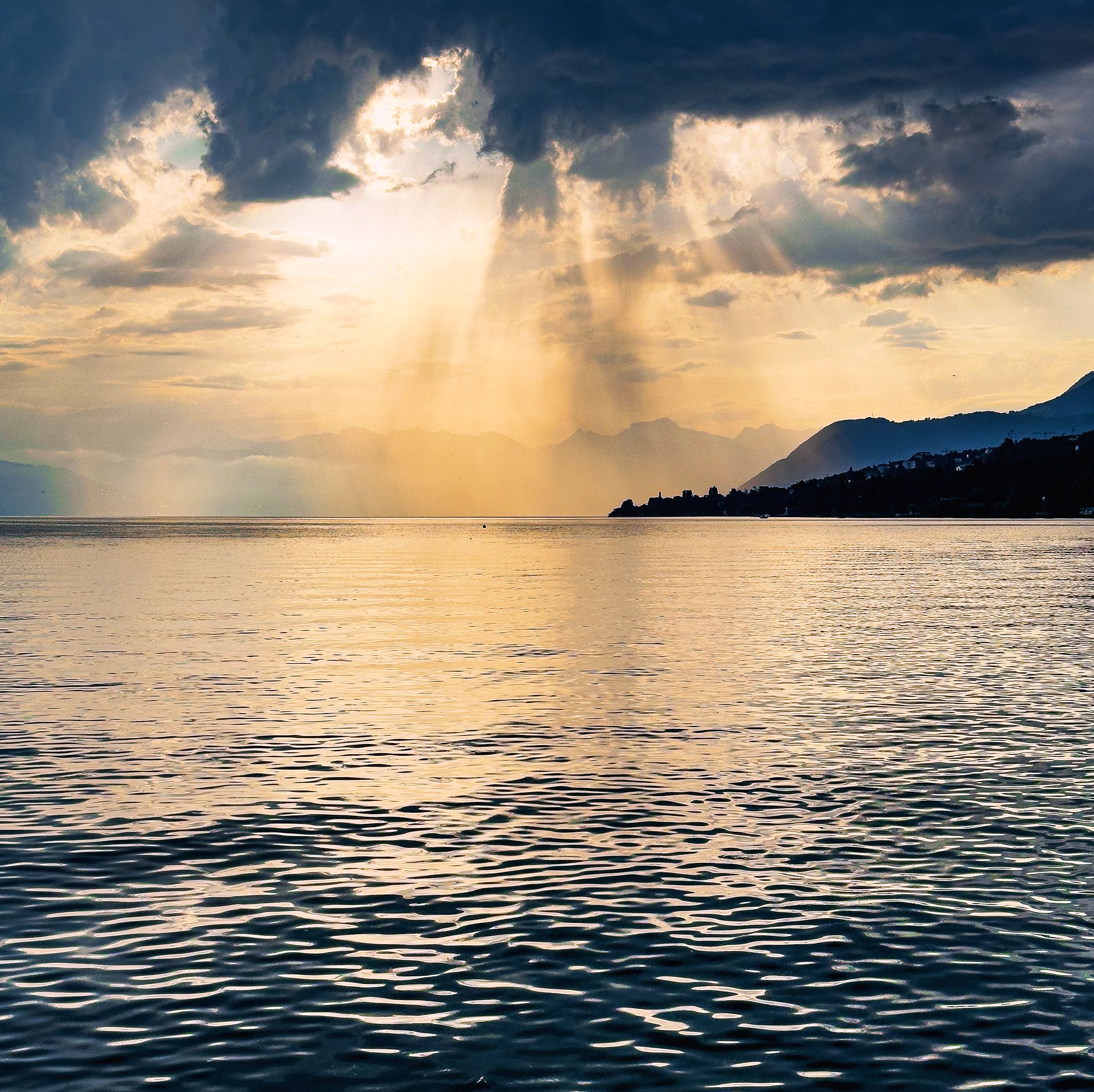 Une mer calme, avec la lumière du soleil se reflétant sur l’eau, une chaîne de montagnes lointaine et un ciel nuageux.