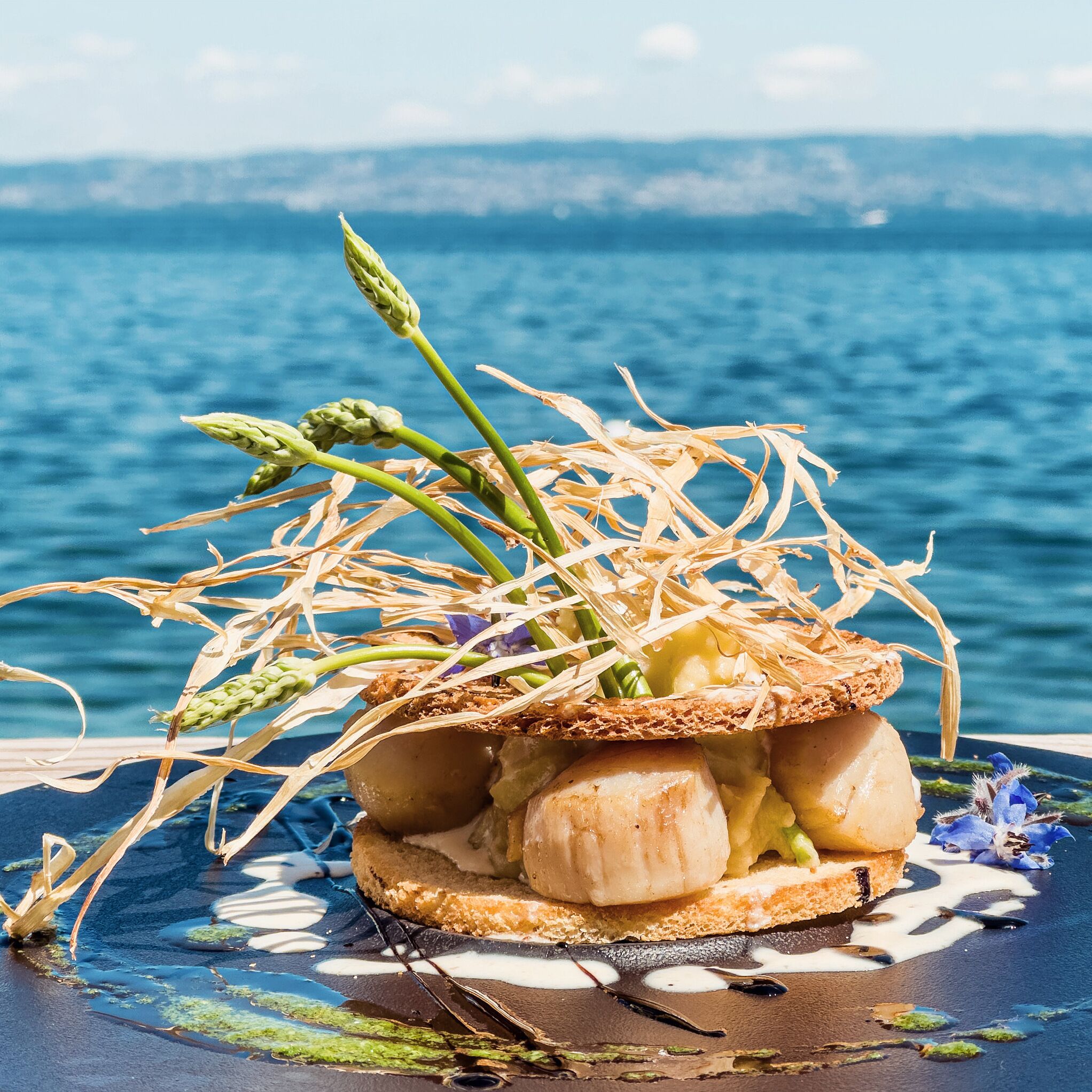 A dessert with a bread base, topped with a creamy substance and garnished with asparagus and flowers, set against a scenic backdrop of water and sky.