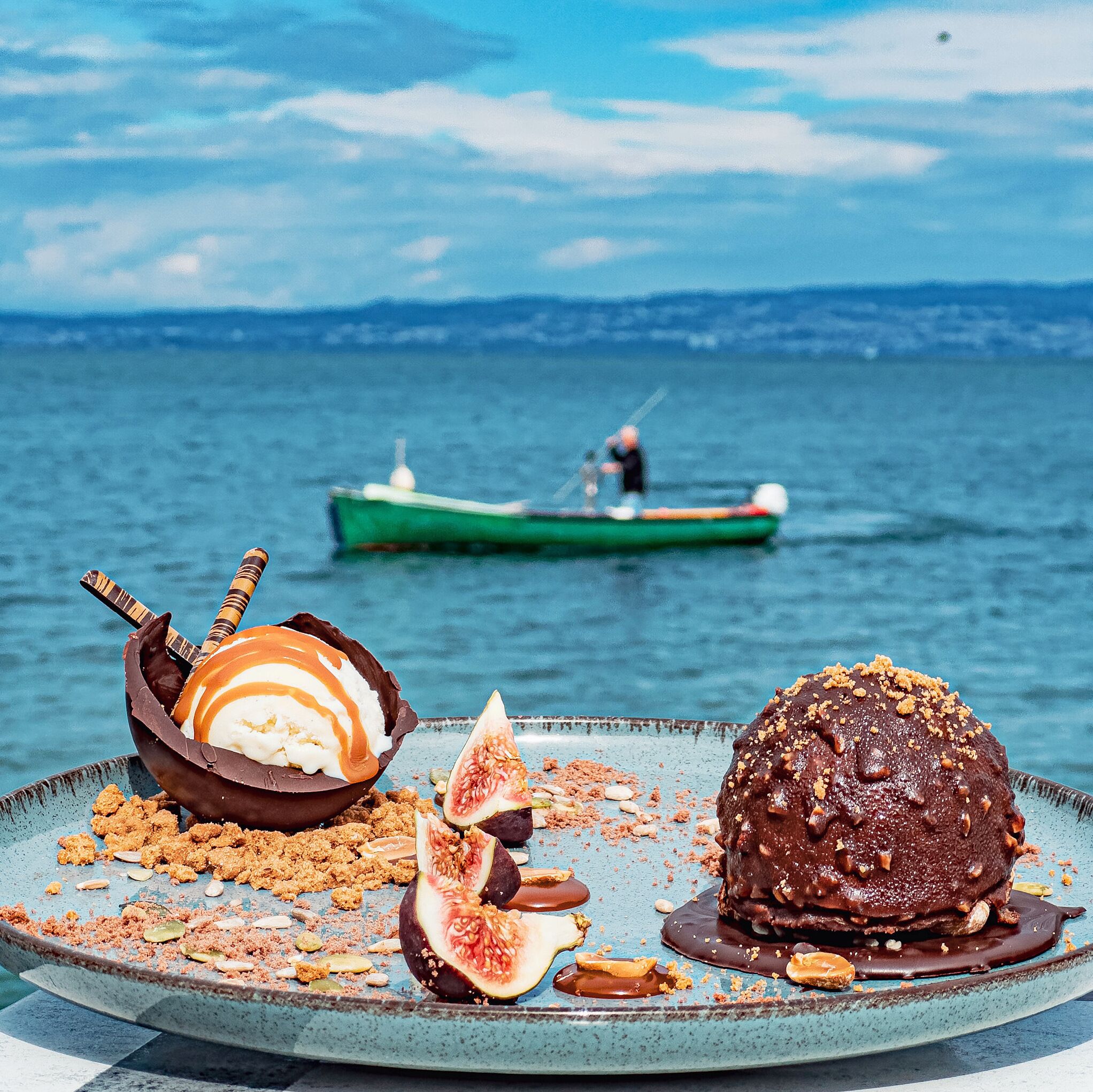 A dessert plate with chocolate and caramel, figs, and nuts on a blue plate by a lake with a boat in the background.