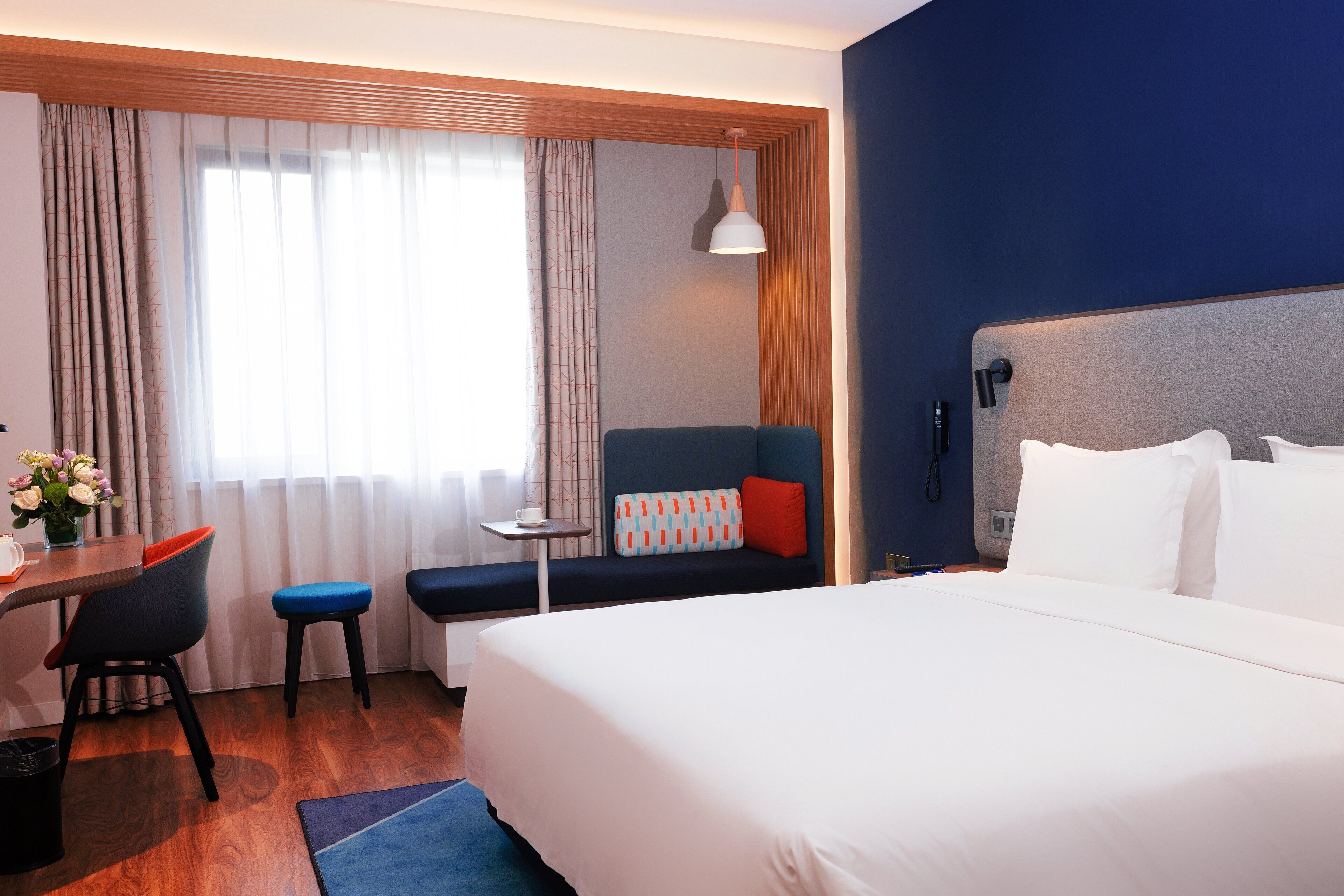 Habitación de hotel con una cama grande, un escritorio con silla, una mesita pequeña y un sofá con un cojín rojo.