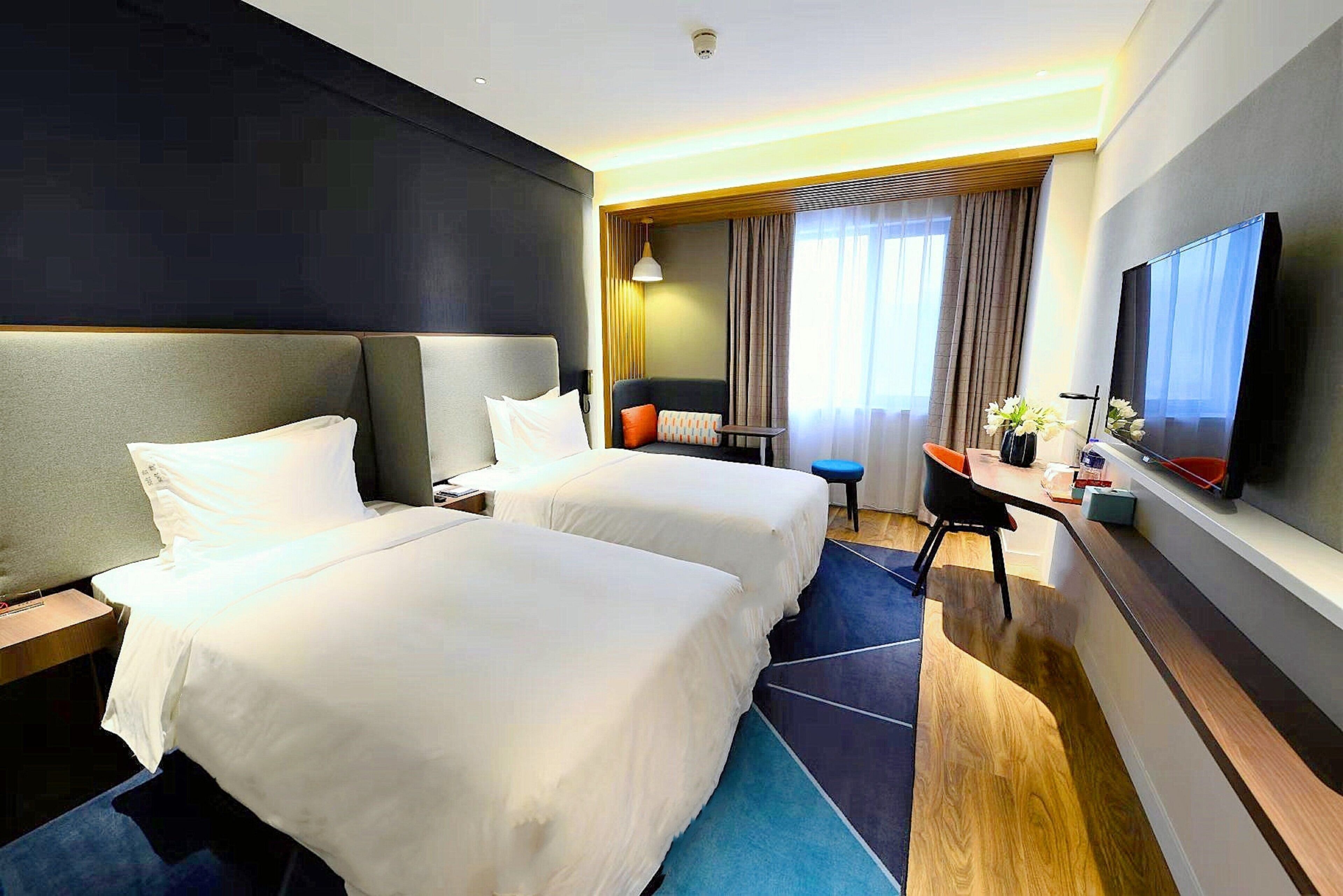 A hotel room with two beds, a desk, a chair, a television, and a large window with curtains.
