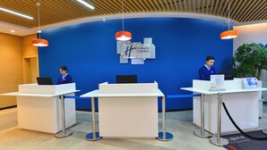 Reseptiotiskialue, jossa on sininen seinä ja kyltti, jossa lukee ”Holiday Inn Express”. Tiskien takana istuu kolme vastaanottovirkailijaa, ja oikealla puolella näkyy ruukussa oleva kasvi.