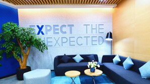 Un hall d’entrée moderne aux couleurs bleu et gris, comprenant un canapé, deux tables rondes, un arbre en pot et un mur sur lequel est inscrit le texte : « ATTENDEZ-VOUS À CE QUI EST ATTENDU ».