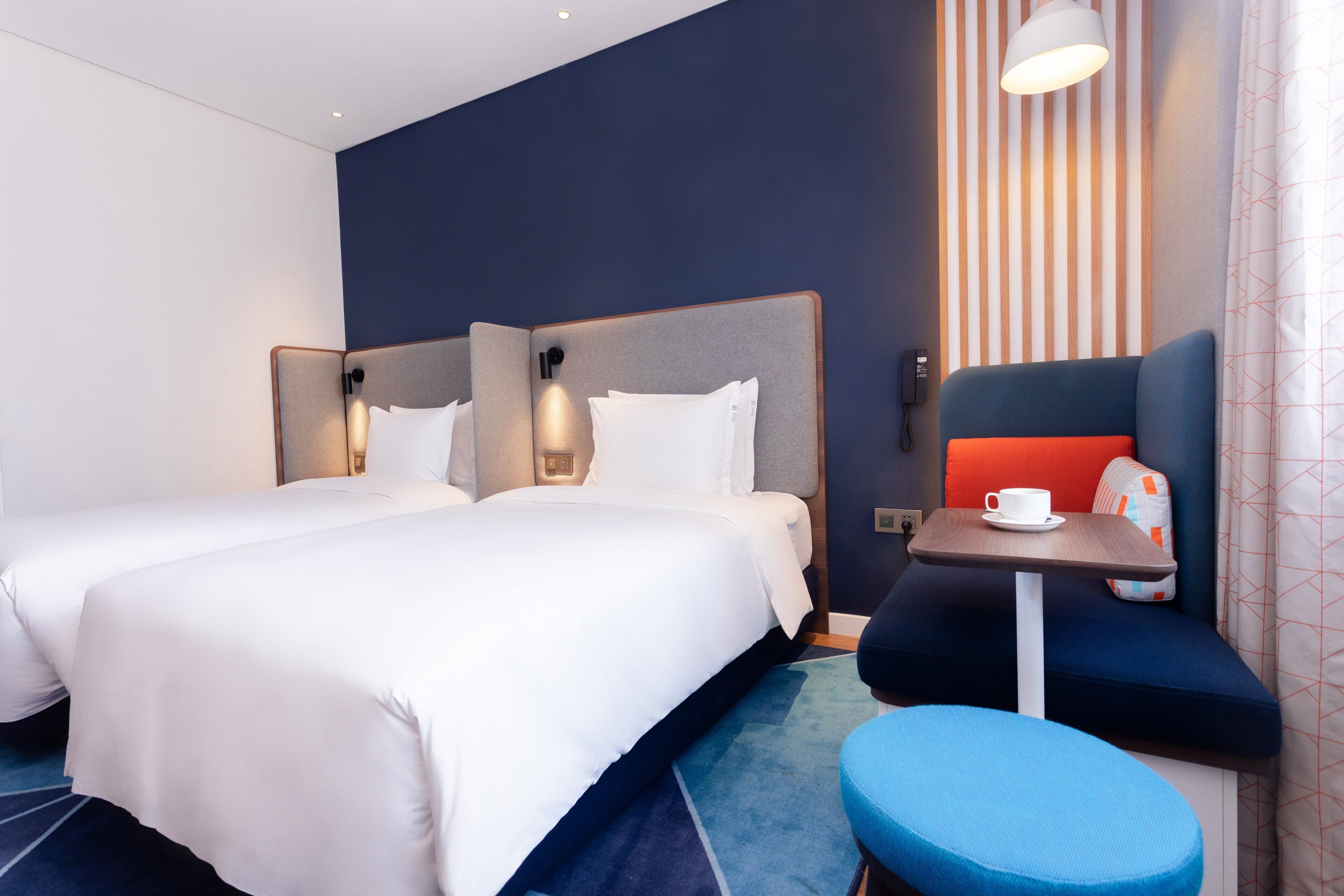 Quarto de hotel com duas camas, uma mesinha com uma chávena e um banco azul.