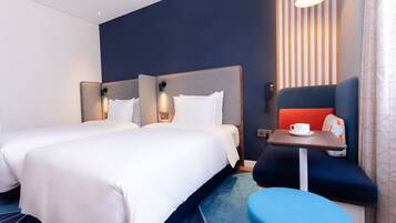 Quarto de hotel com duas camas, uma mesinha com uma chávena e um banco azul.