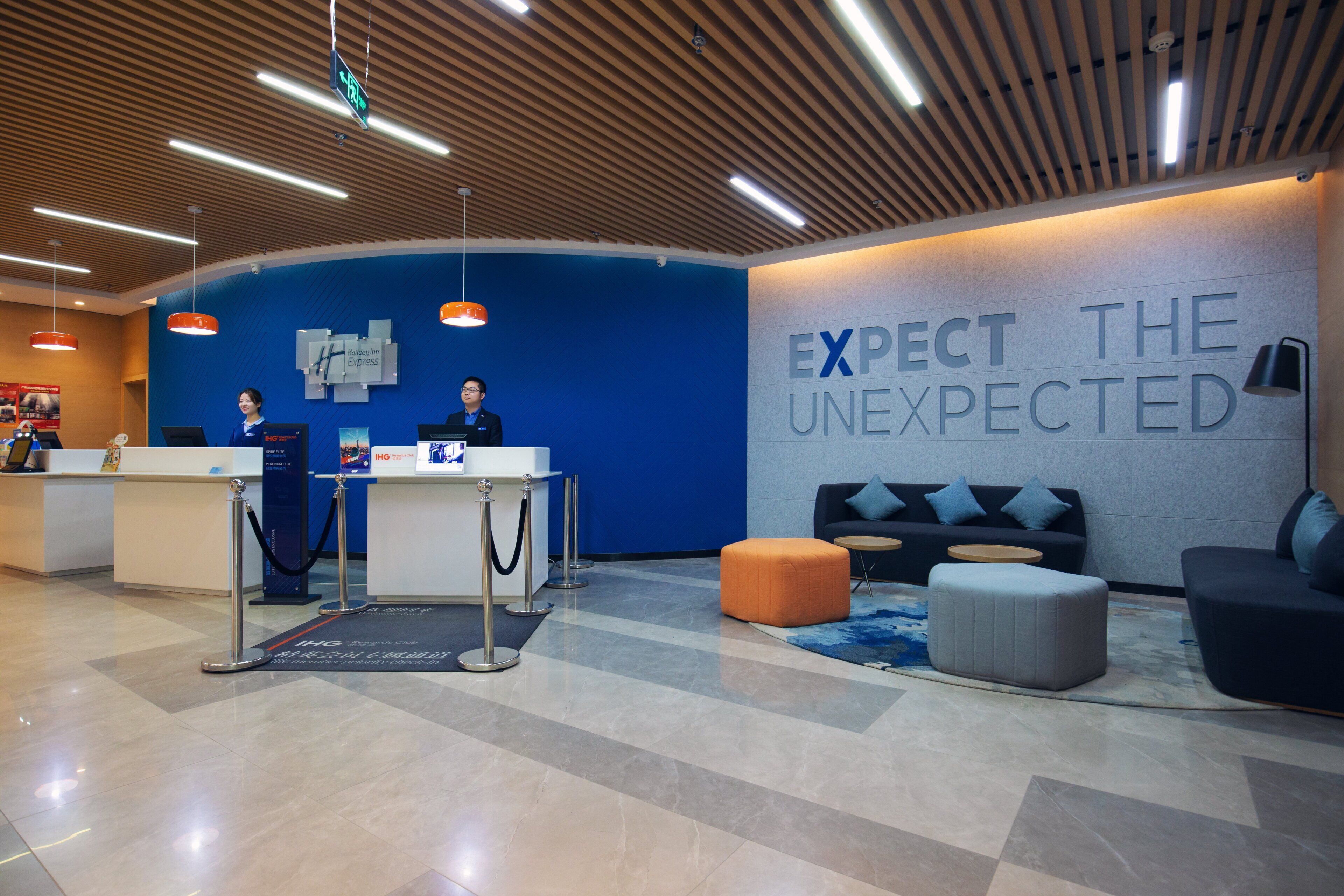 Zona de recepción de hotel con pared azul, un cartel que dice 'EXPECT THE UNEXPECTED' y un mostrador de recepción con dos empleados.
