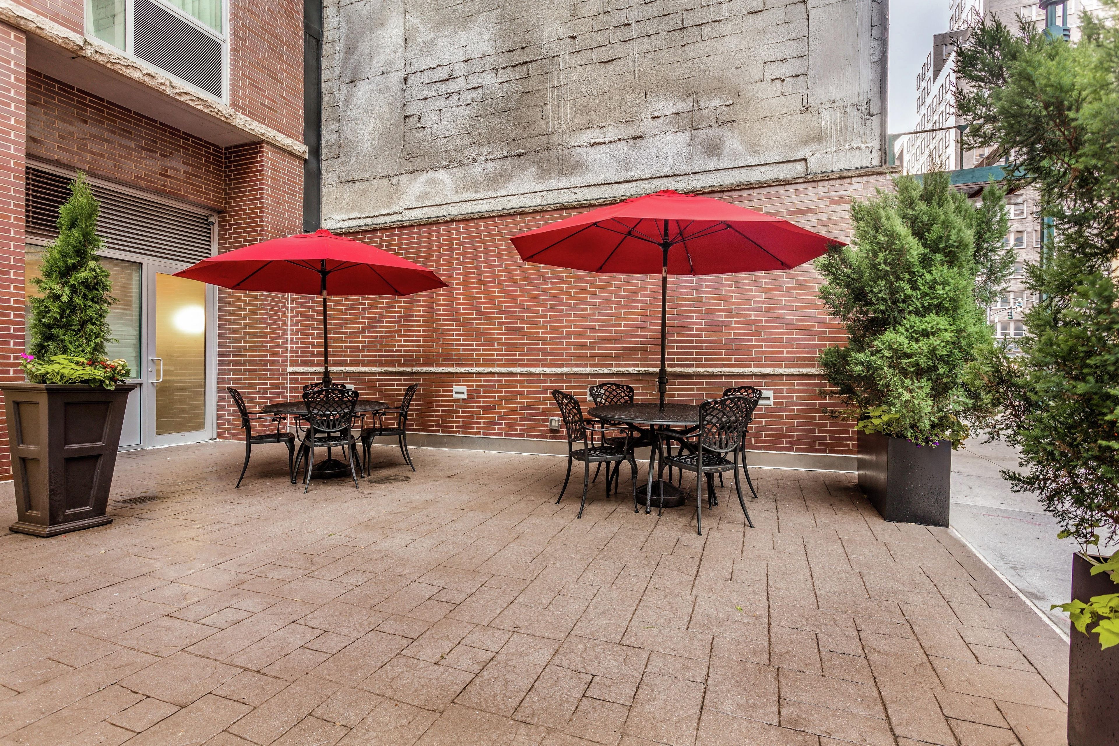 An outdoor patio with brick flooring, two red umbrellas, and metal tables and chairs.