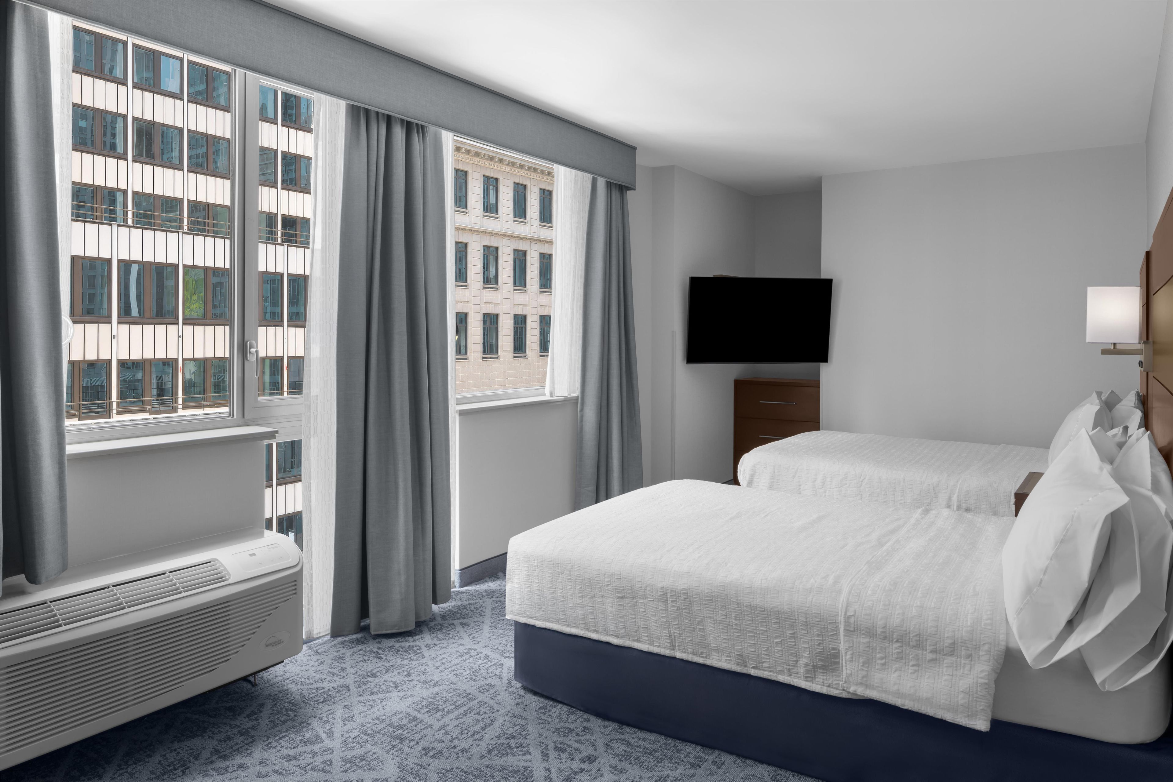 Habitación de hotel con una ventana grande, televisor de pantalla plana, cama con ropa blanca y una mesita de noche.