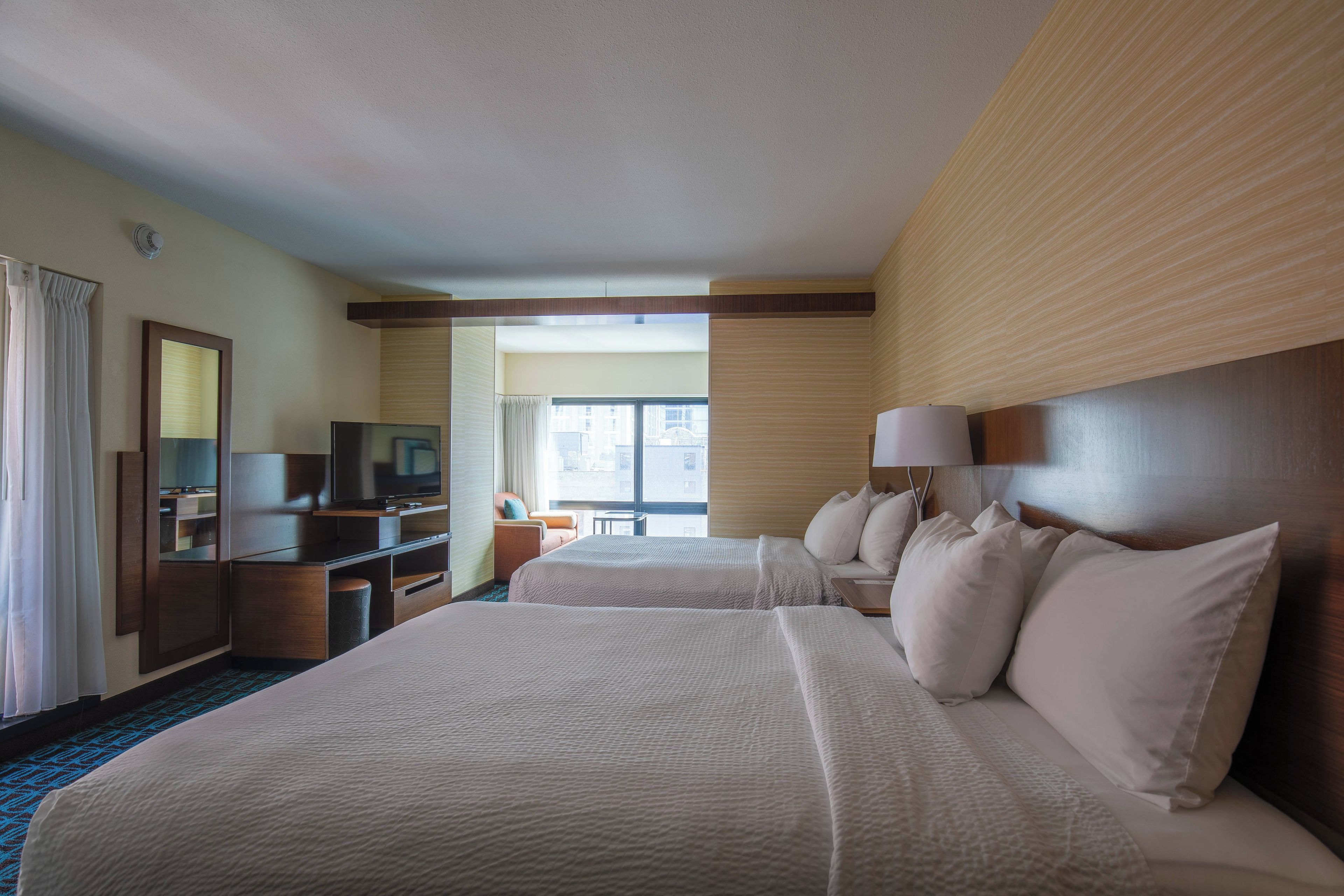 A hotel room with two beds, a television, a desk, and a window with a view of buildings.