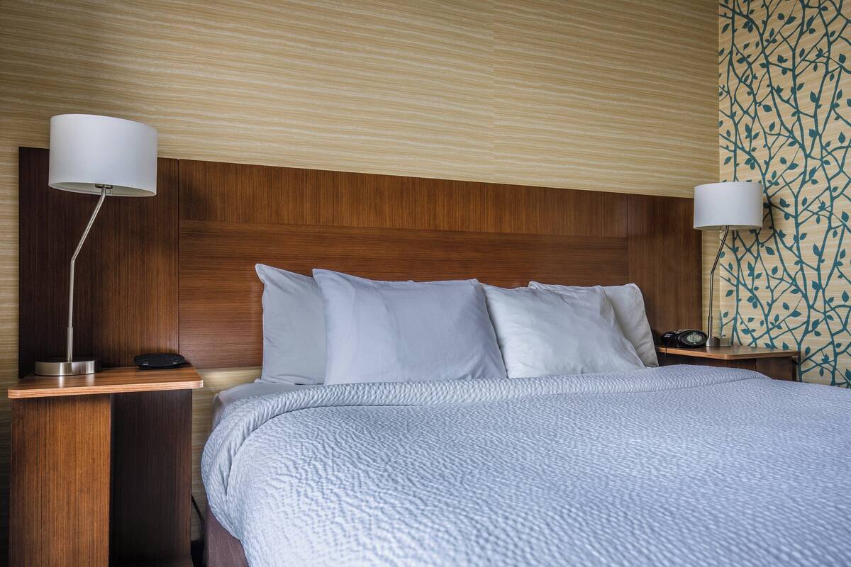 Hotellihuone, jossa on puinen sängynpääty, valkoisilla vuodevaatteilla varustettu sänky, kaksi yöpöytää, joissa on lamput, ja seinä, jossa on lehtimäinen kuvio.