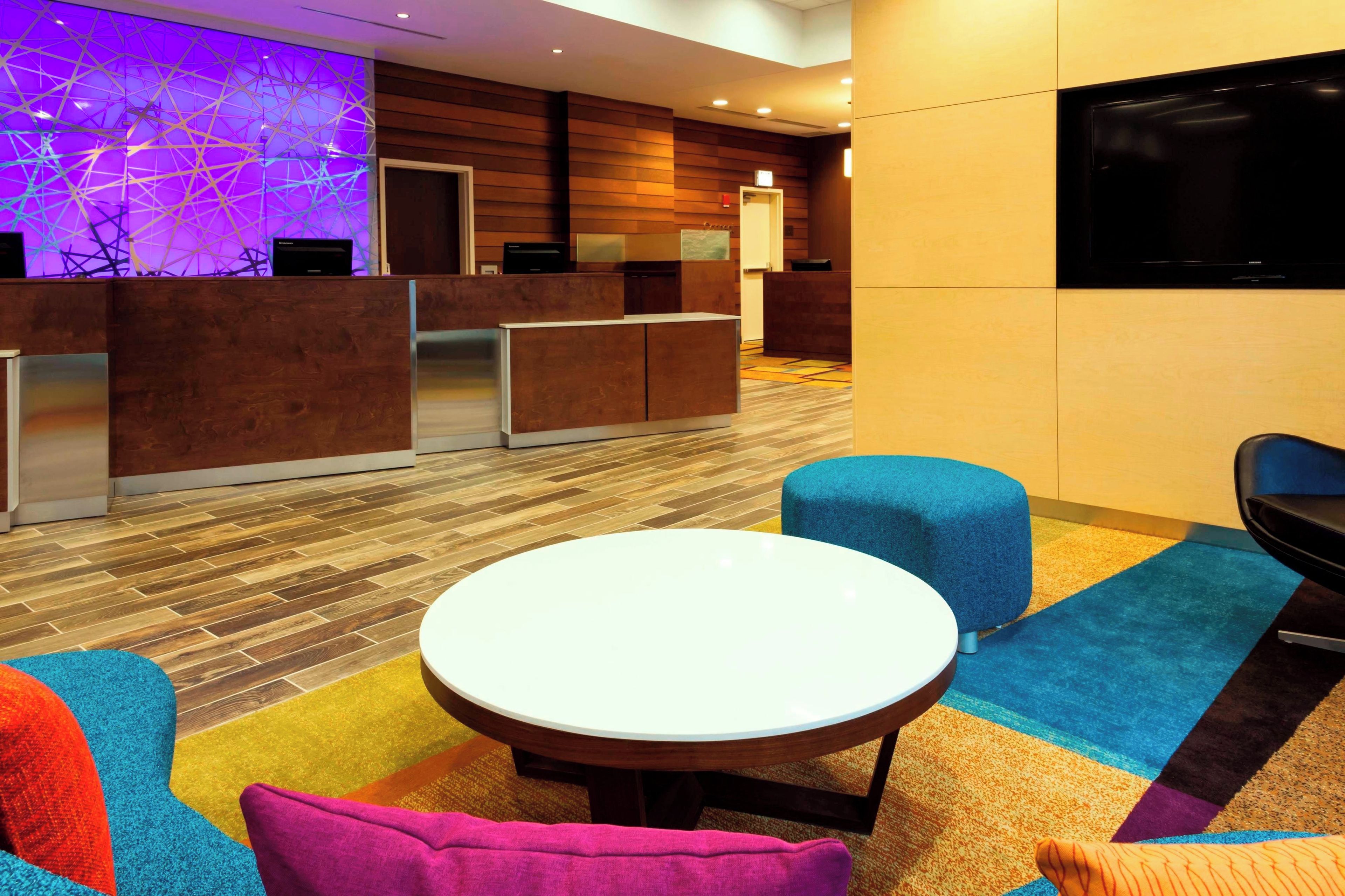 A hotel lobby with a reception desk, a television, a round table, and colorful seating.