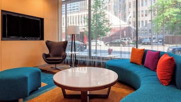A modern hotel room with a large window, a flat-screen TV, a round coffee table, a blue sofa, and a black armchair.